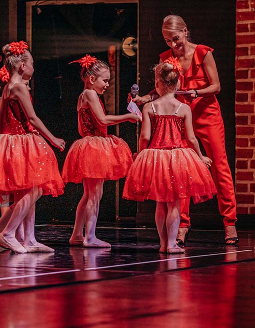 Grupa najmłodszych baletnic w czerwonych tutu na scenie, obok nauczycielka w czerwonym stroju