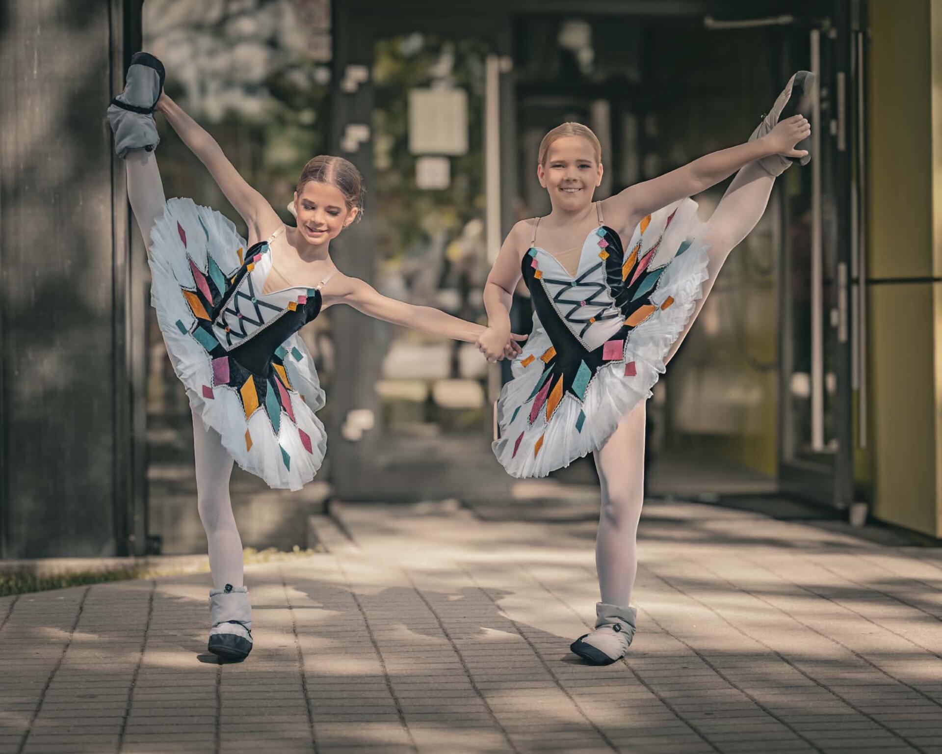 Dwie uczennice w białych tutu w wysokim wyciągnięciu nogi, na chodniku przed budynkiem